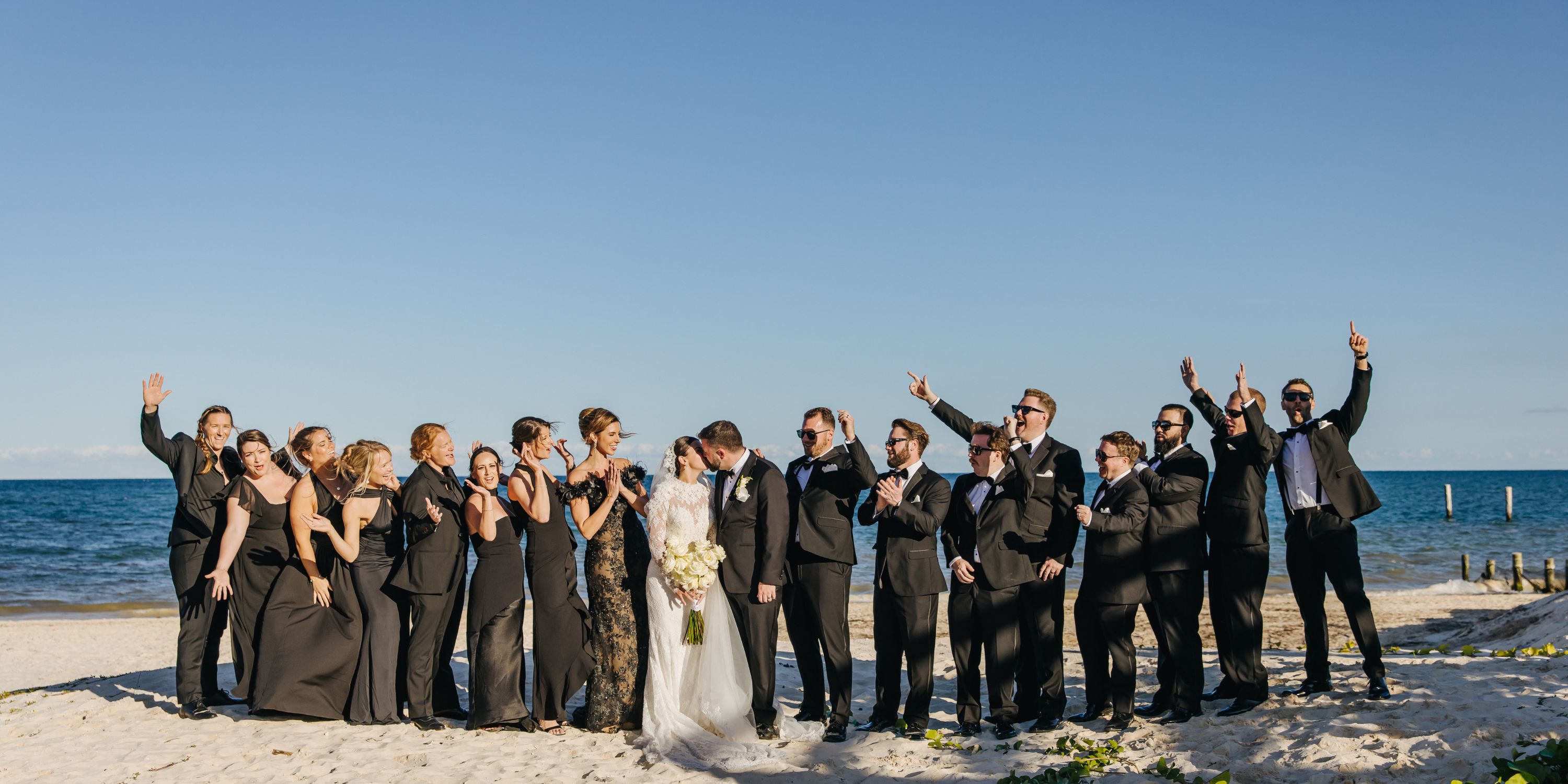 Kathryn and Mike&rsquo;s destination wedding party stands on a sandy beach with ocean and blue sky behind.