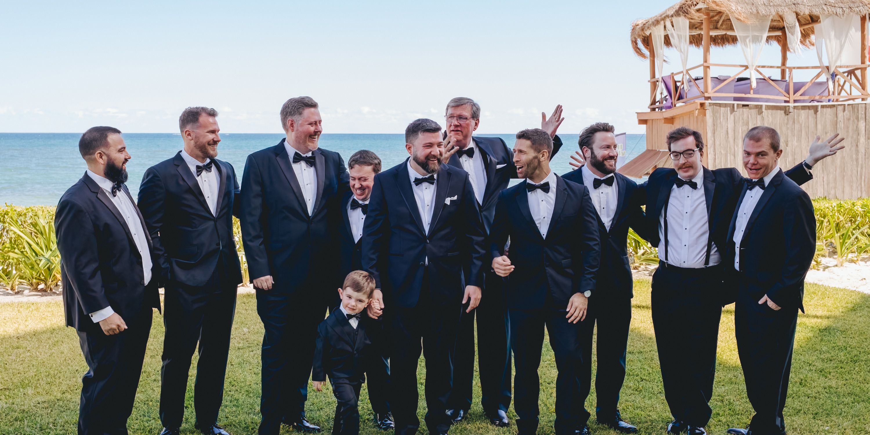 Men and a boy in tuxedos smile outdoors by the ocean at a destination wedding in "Kathryn and Mike (Copy)".