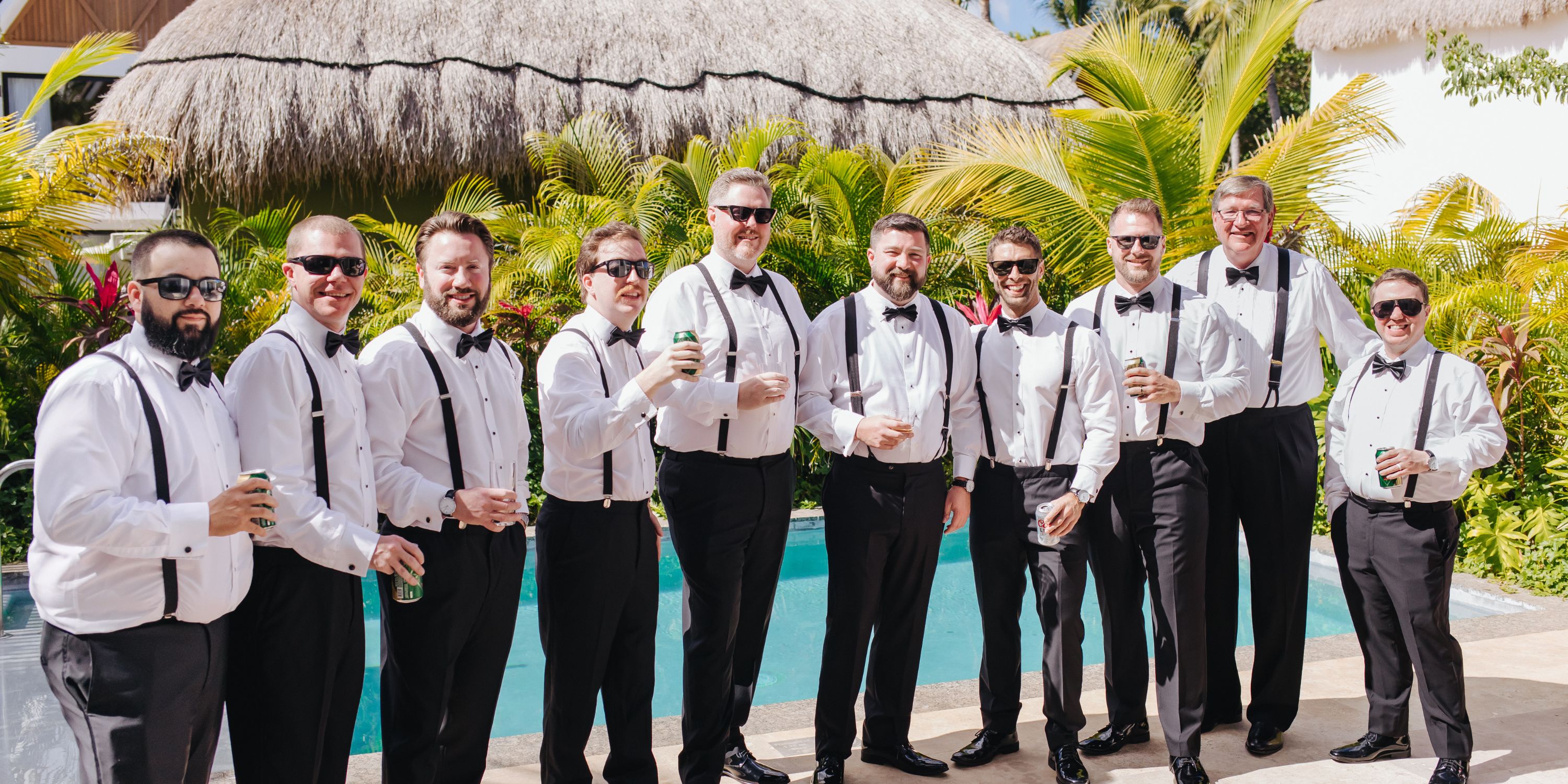 Ten men in suspenders by a pool toast drinks and smile for Kathryn and Mike&rsquo;s destination wedding.