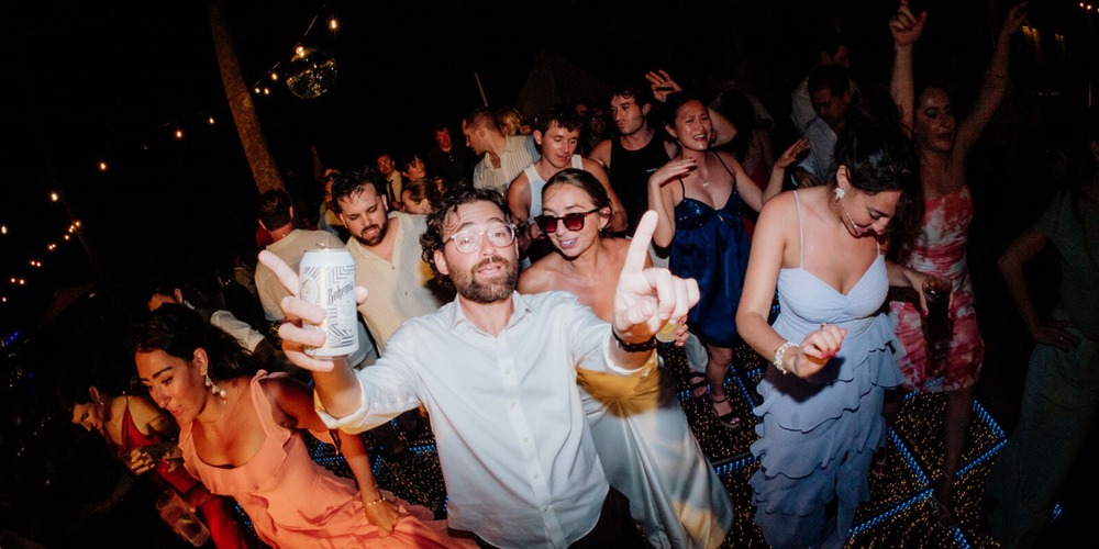 Guests dance at a destination wedding; one man with a drink points at the camera. Ethan and Erica.