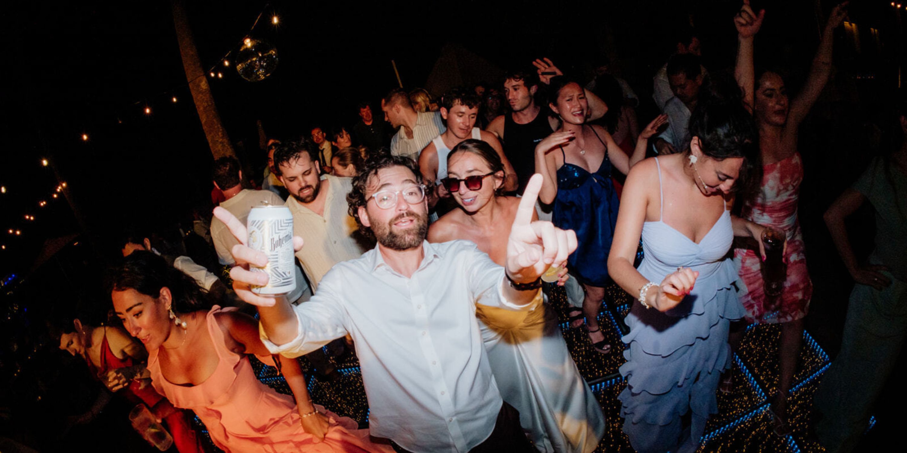 Guests dance at a destination wedding; one man with a drink points at the camera. Ethan and Erica.