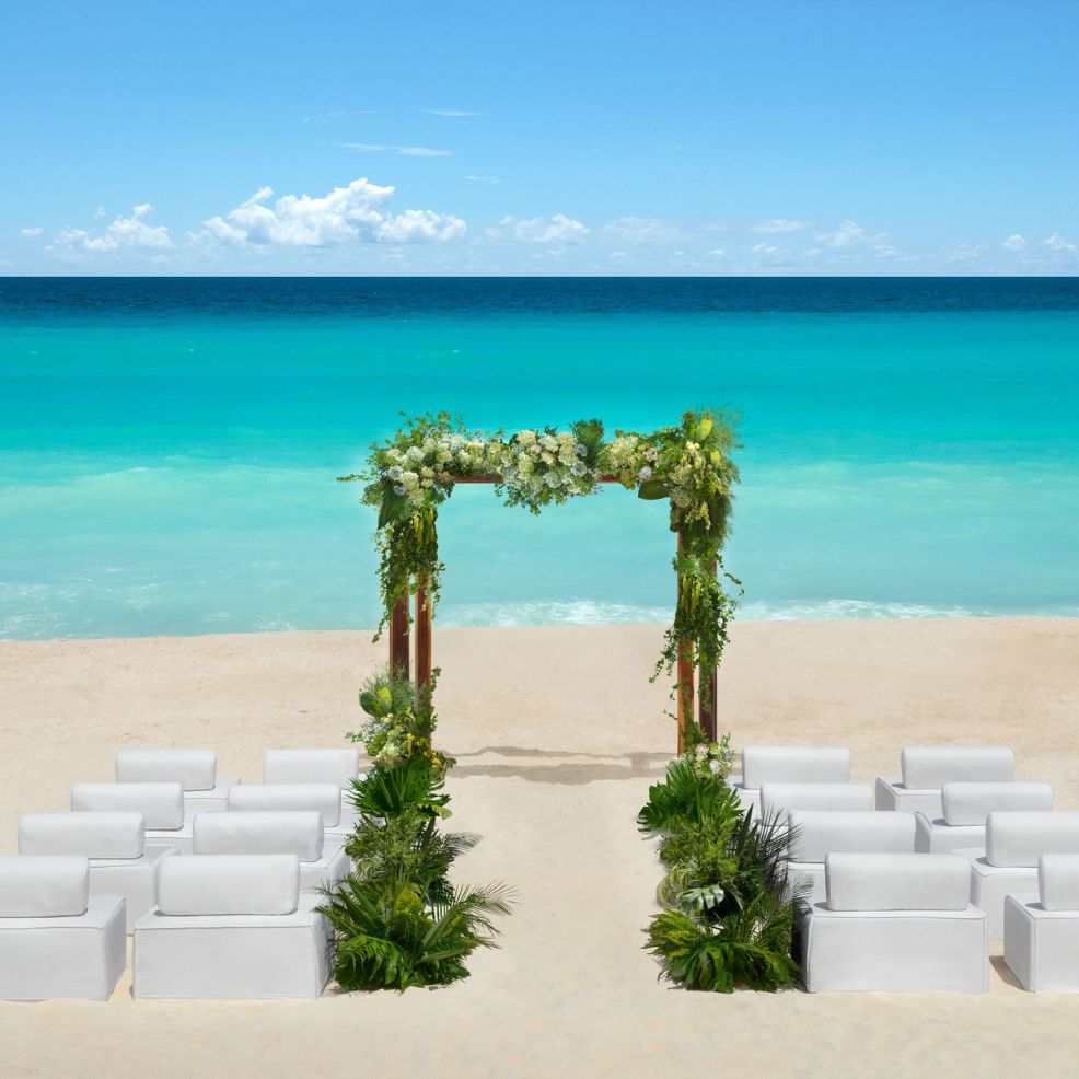 A destination wedding setup with a white arch and chairs on sandy beach overlooking the turquoise ocean.