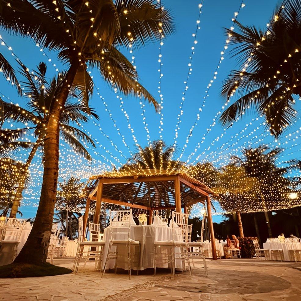 Destination wedding setup beneath palm trees at dusk, with string lights and white-clothed tables.