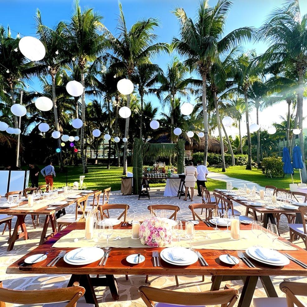 Destination wedding setup featuring wooden tables, white plates, and lanterns hanging beneath palm trees.