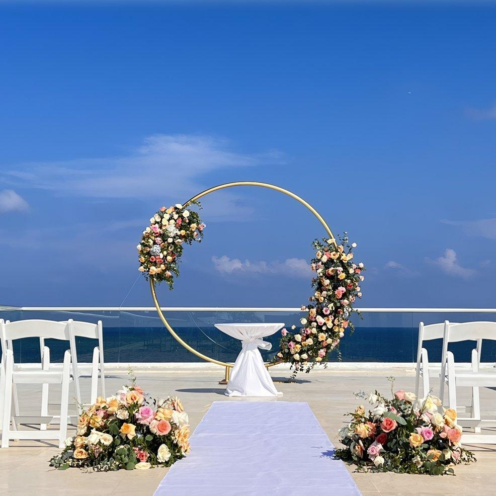 Destination wedding ceremony featuring a round floral arch, white chairs, and an ocean view under blue sky.