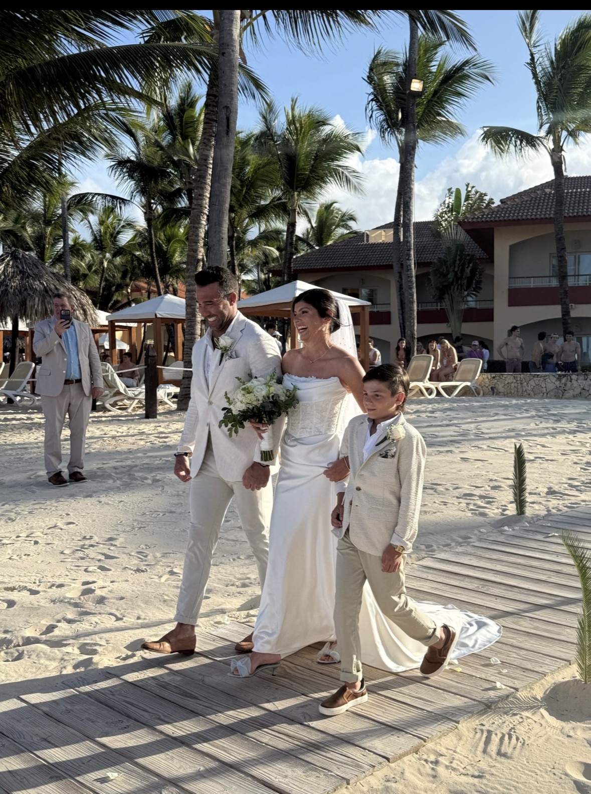 Bride, groom, and boy stroll a sandy boardwalk at a tropical resort destination wedding.