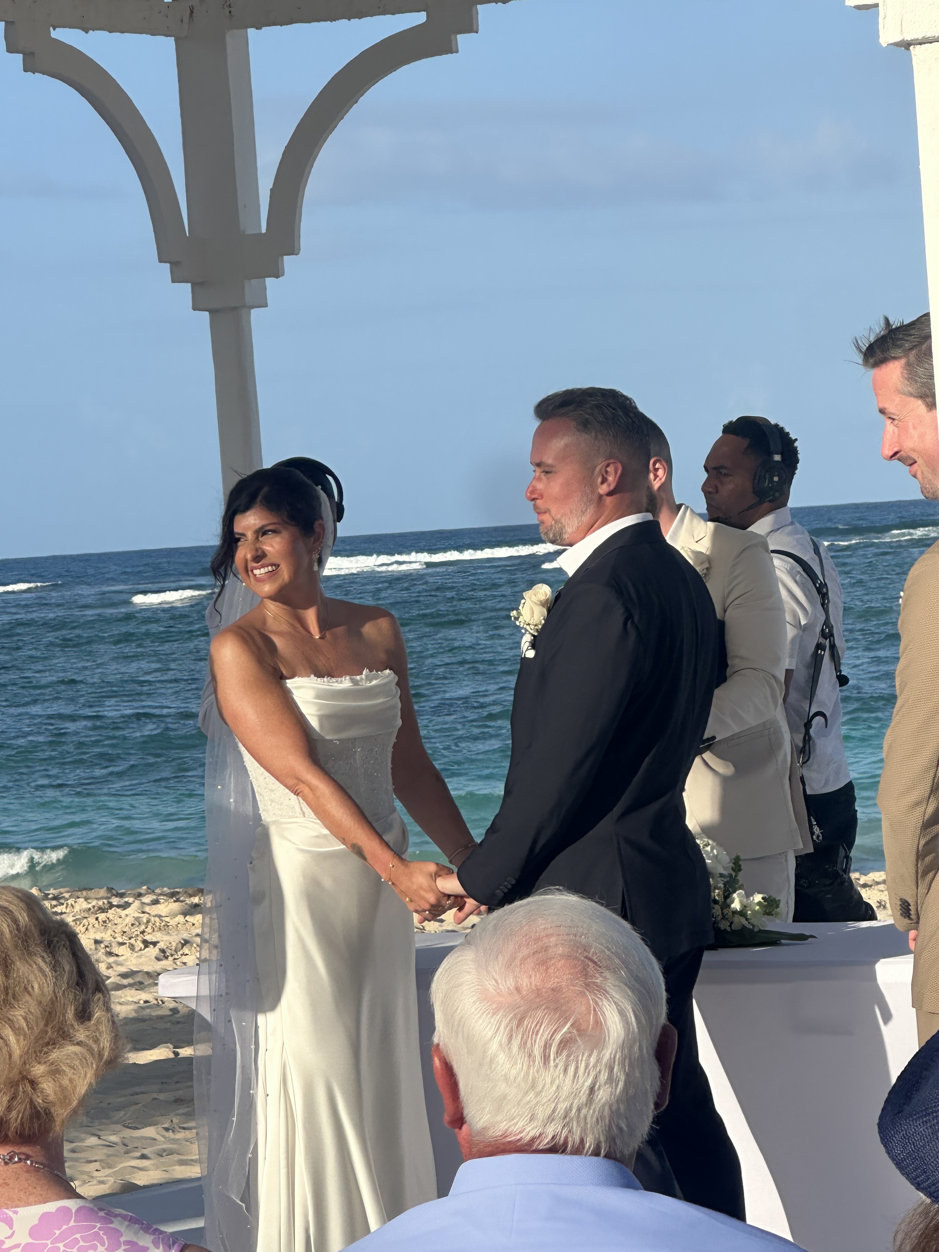 Hannah and Ben hold hands during their destination wedding under a white gazebo on the beach.