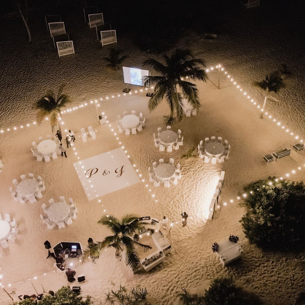 Aerial shot of a destination wedding reception on the beach, round tables and string lights glowing at night.