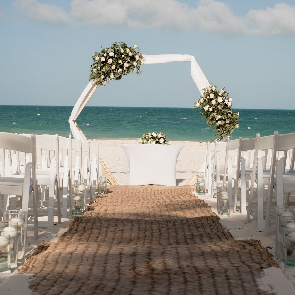 Destination wedding scene with white chairs, floral arch, and woven aisle arranged toward the ocean.