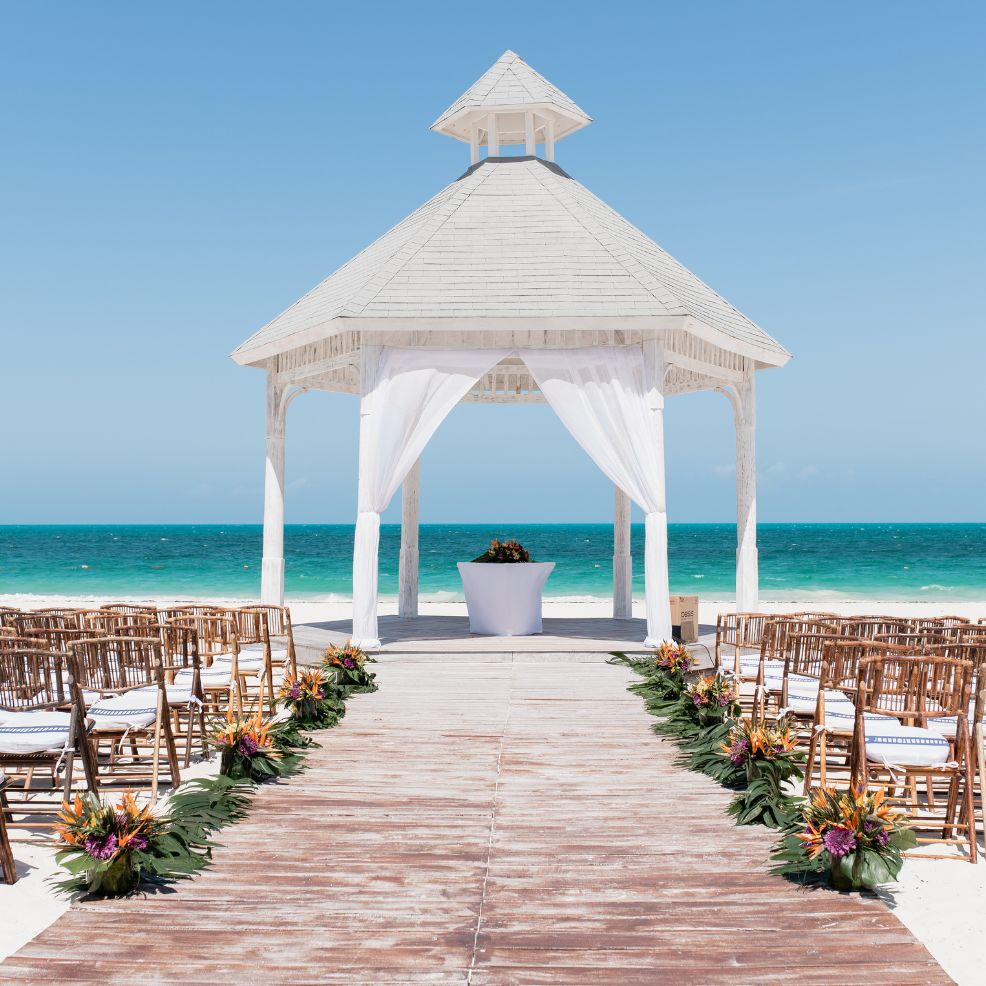 A white gazebo on a beach set up for a destination wedding, with chairs and flowers ready for guests.