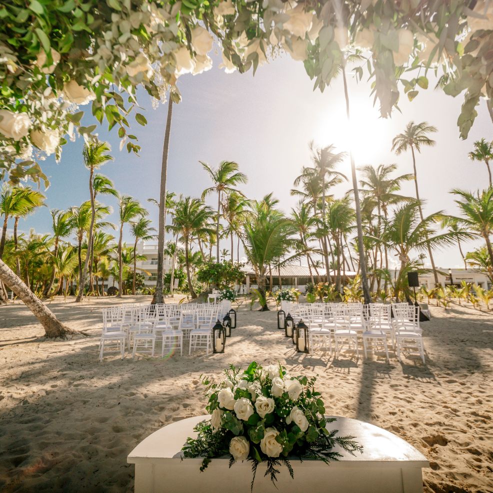Chairs and flowers arranged for a destination wedding on the beach, with palm trees in bright sunlight.