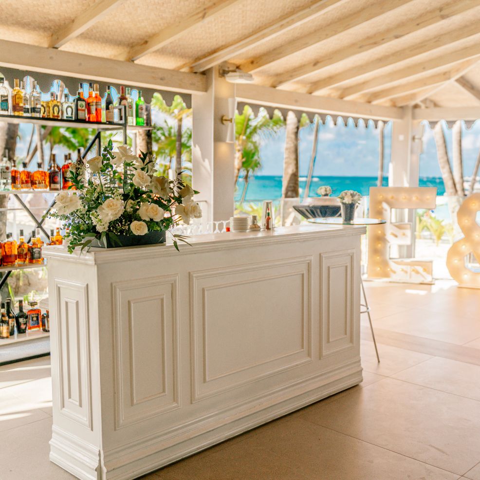White bar counter with floral d&eacute;cor, liquor shelves, and a beach view perfect for a destination wedding.