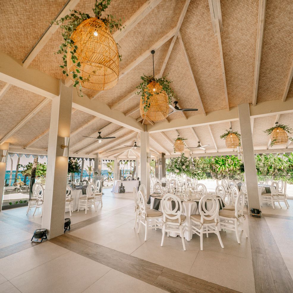 Open-air wedding dining area with round tables, white chairs, and hanging wicker lamps.