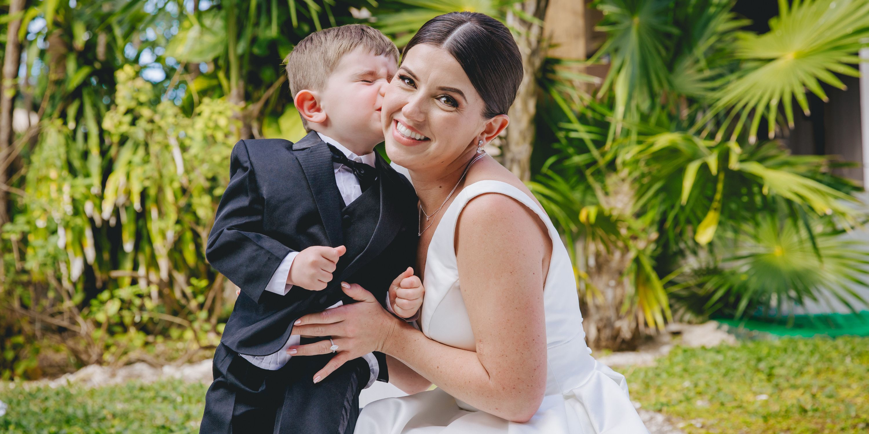 Young boy in a tuxedo kisses smiling woman in white dress by greenery at a destination wedding.