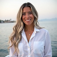Woman with long blonde hair in a white shirt by the water, smiling as if at a destination wedding.