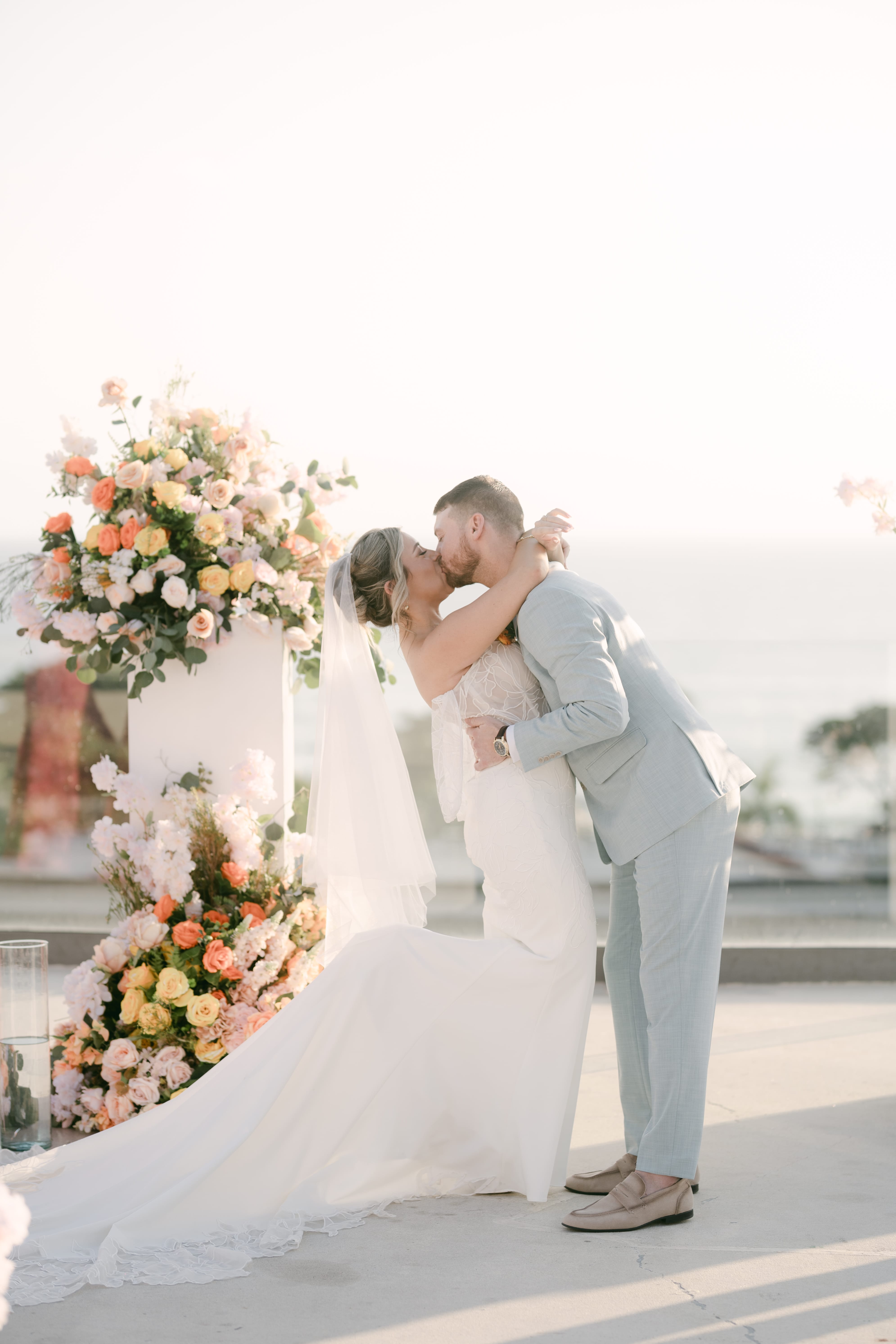 Bride and groom share a kiss at their destination wedding, surrounded by pastel floral arrangements outdoors.