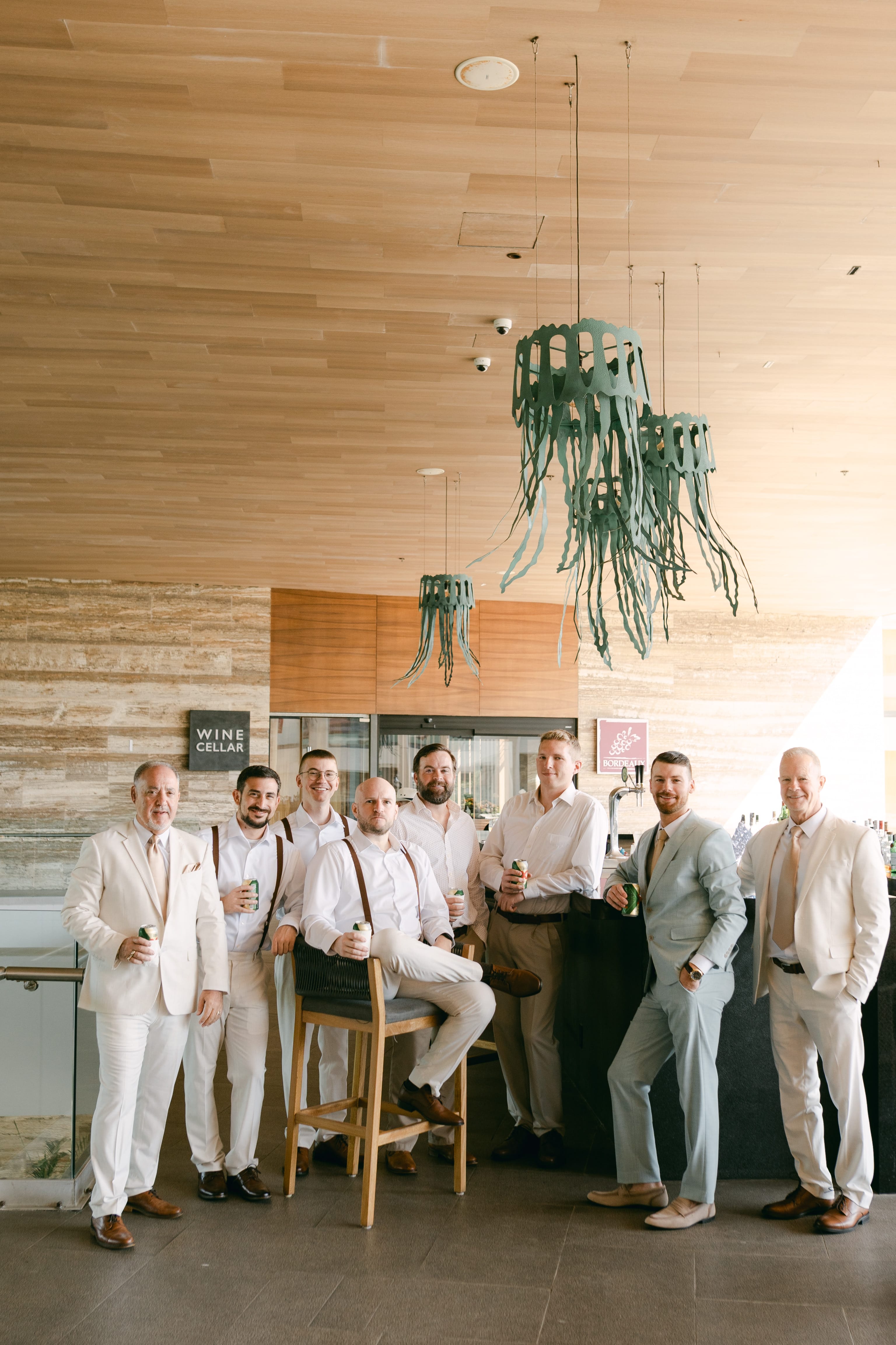 Eight men in light suits pose at a bar under modern lamps during a stylish destination wedding event.