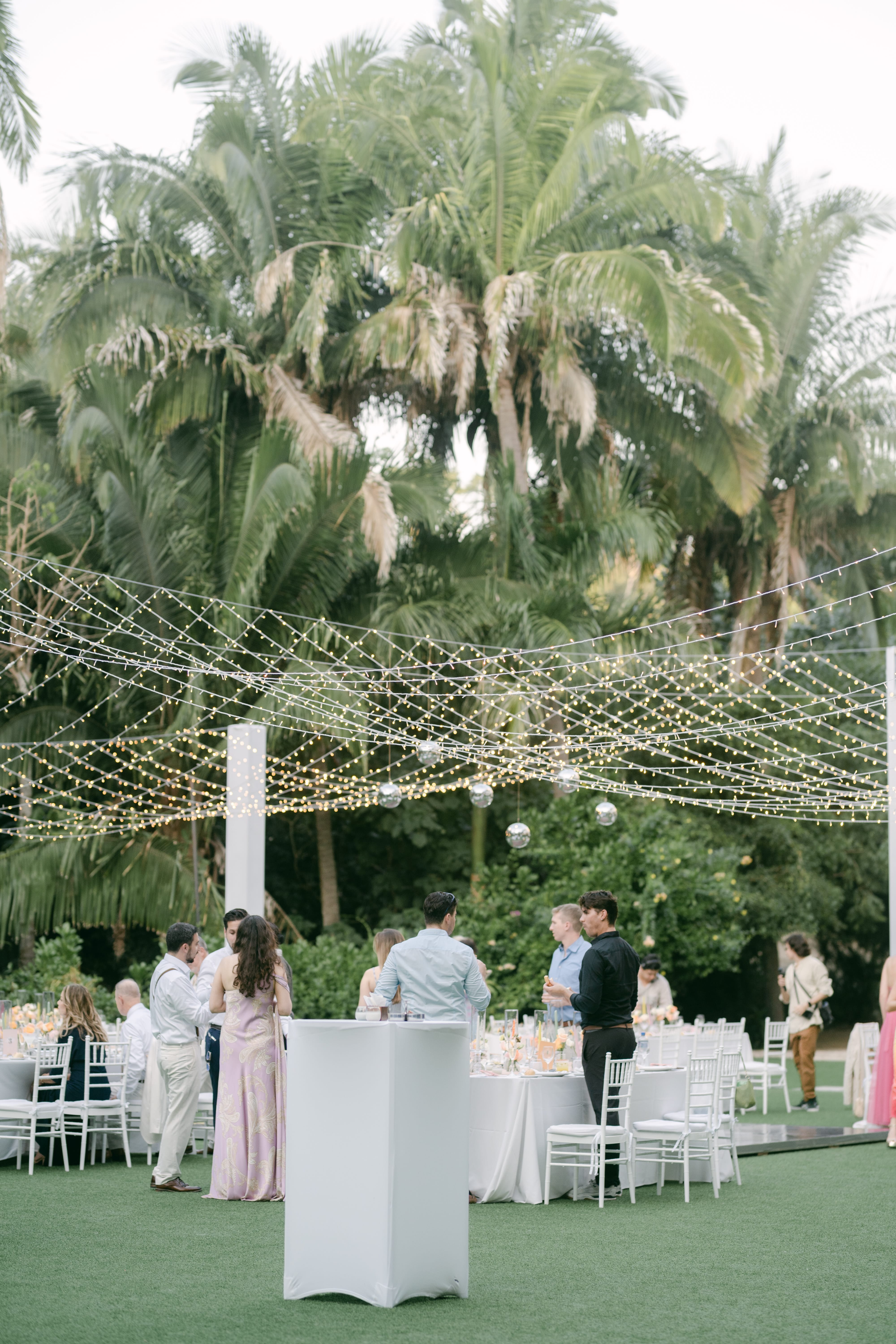 Ashley and Zach enjoy their destination wedding outdoors, seated at round tables under string lights and palm trees.