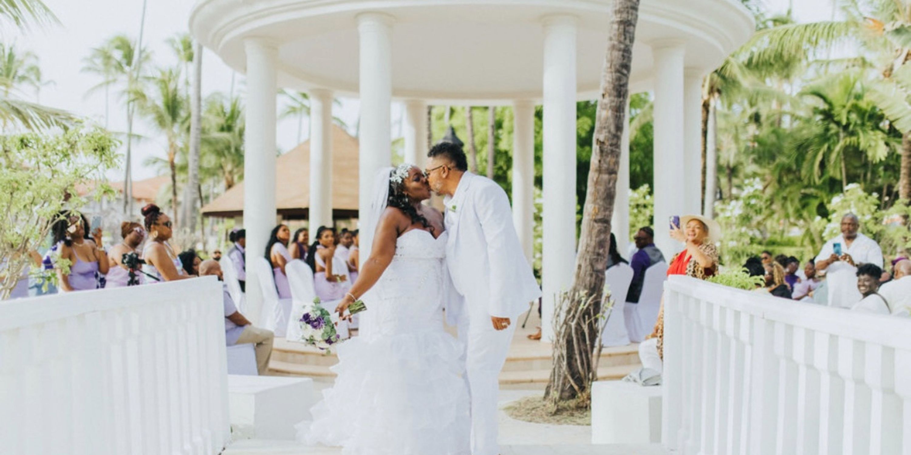 Ethan and Erica share a kiss in white at their destination wedding as guests look on outdoors.
