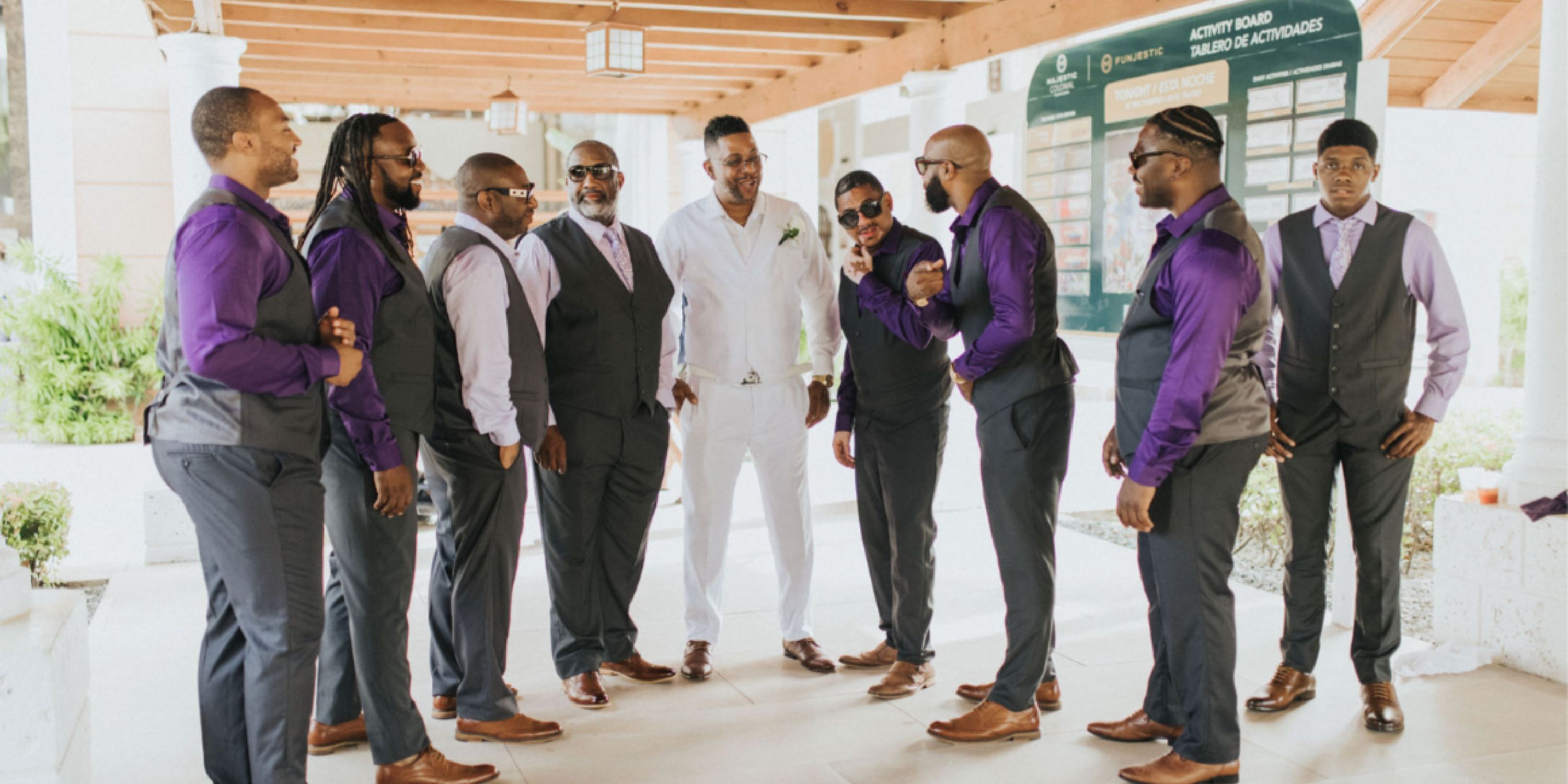 Eight men in Ethan and Erica vests stand indoors at a destination wedding, one all in white.