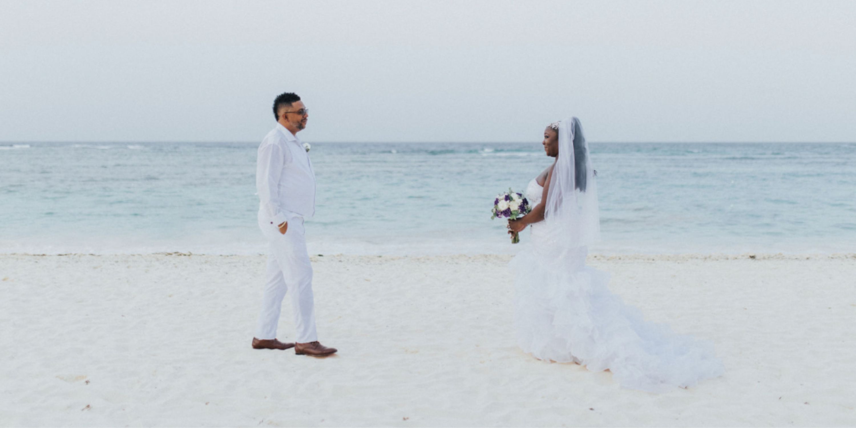 Ethan and Erica face each other on a sandy beach with the ocean behind them at a destination wedding.