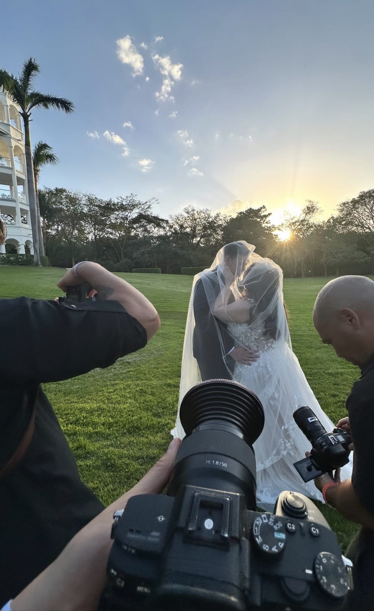 Ashley and Zach kiss beneath a veil at sunset during their destination wedding, captured by photographers.