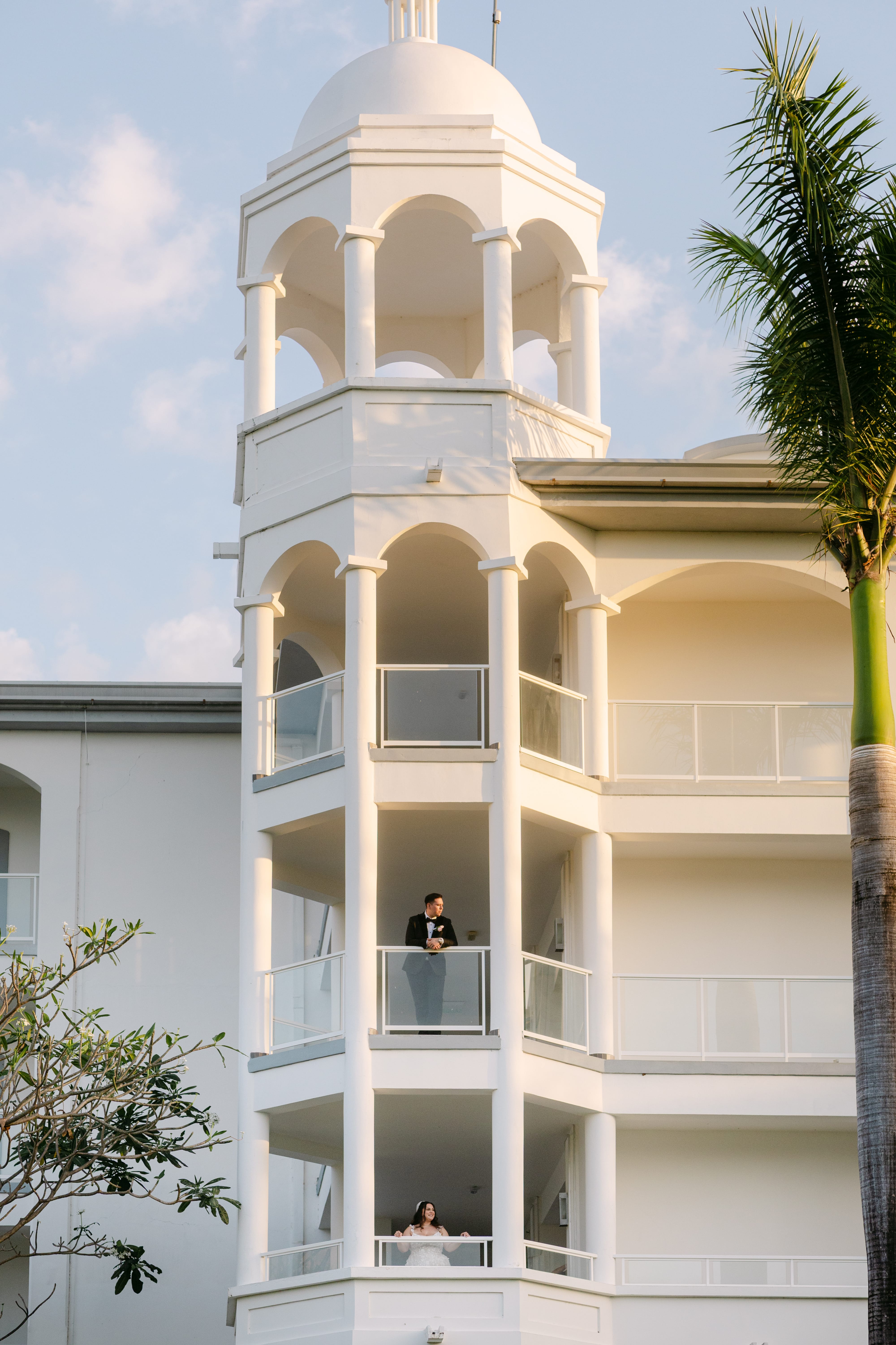 Ashley and Zach stand apart on balconies of a white building at their destination wedding.