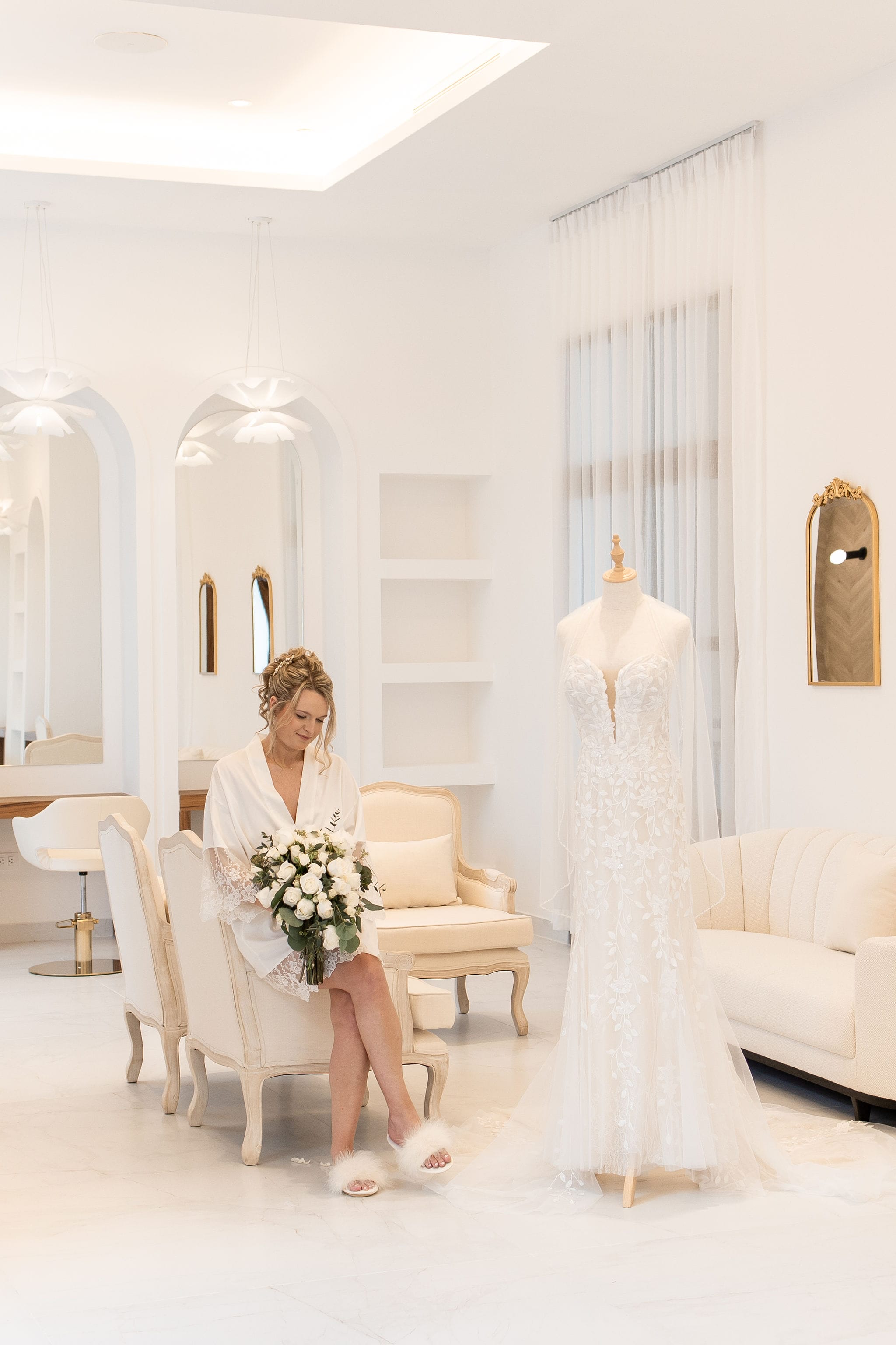 Woman in a white robe holding a bouquet sits next to a Lauren and Ben wedding dress in a bright room, ready for the destination wedding.