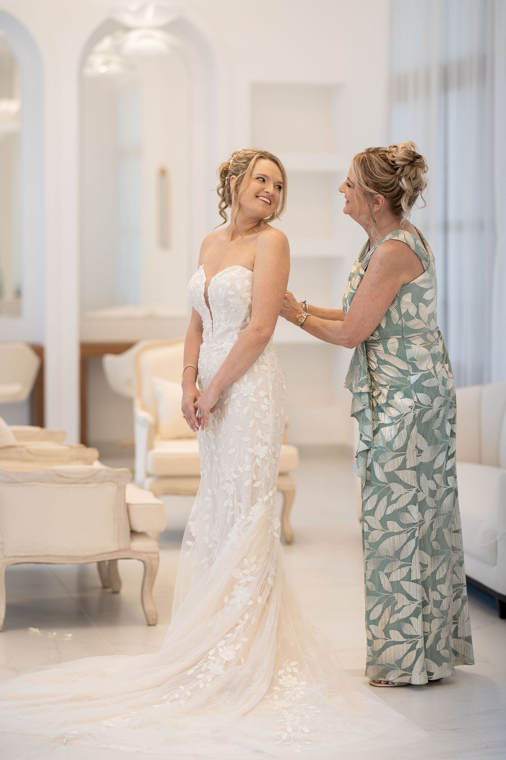 A woman assists a bride with her dress in a bright, elegant room before a destination wedding.