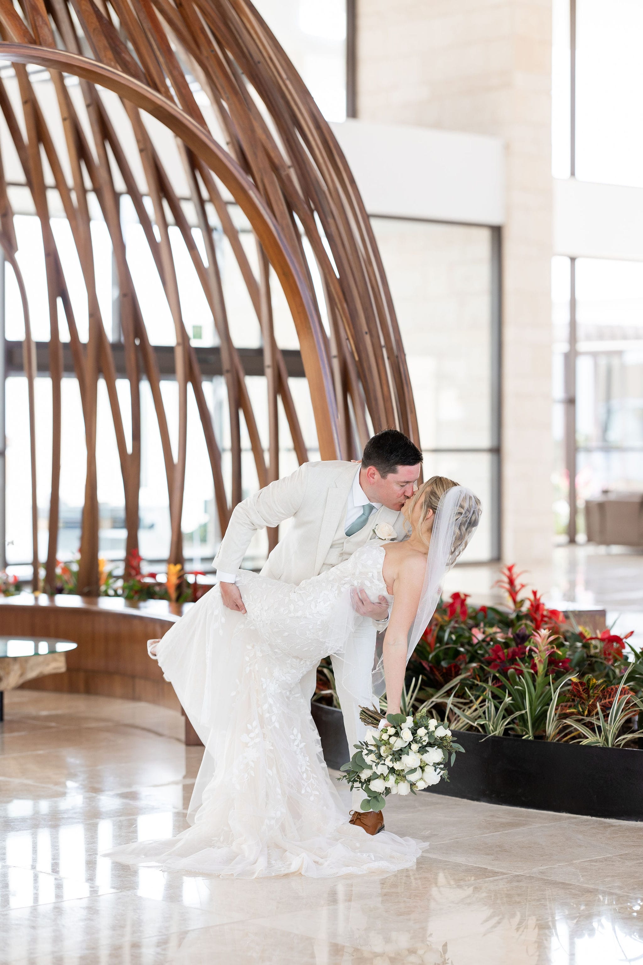 Lauren and Ben share a wedding embrace as the groom kisses the bride in white by lush indoor plants.