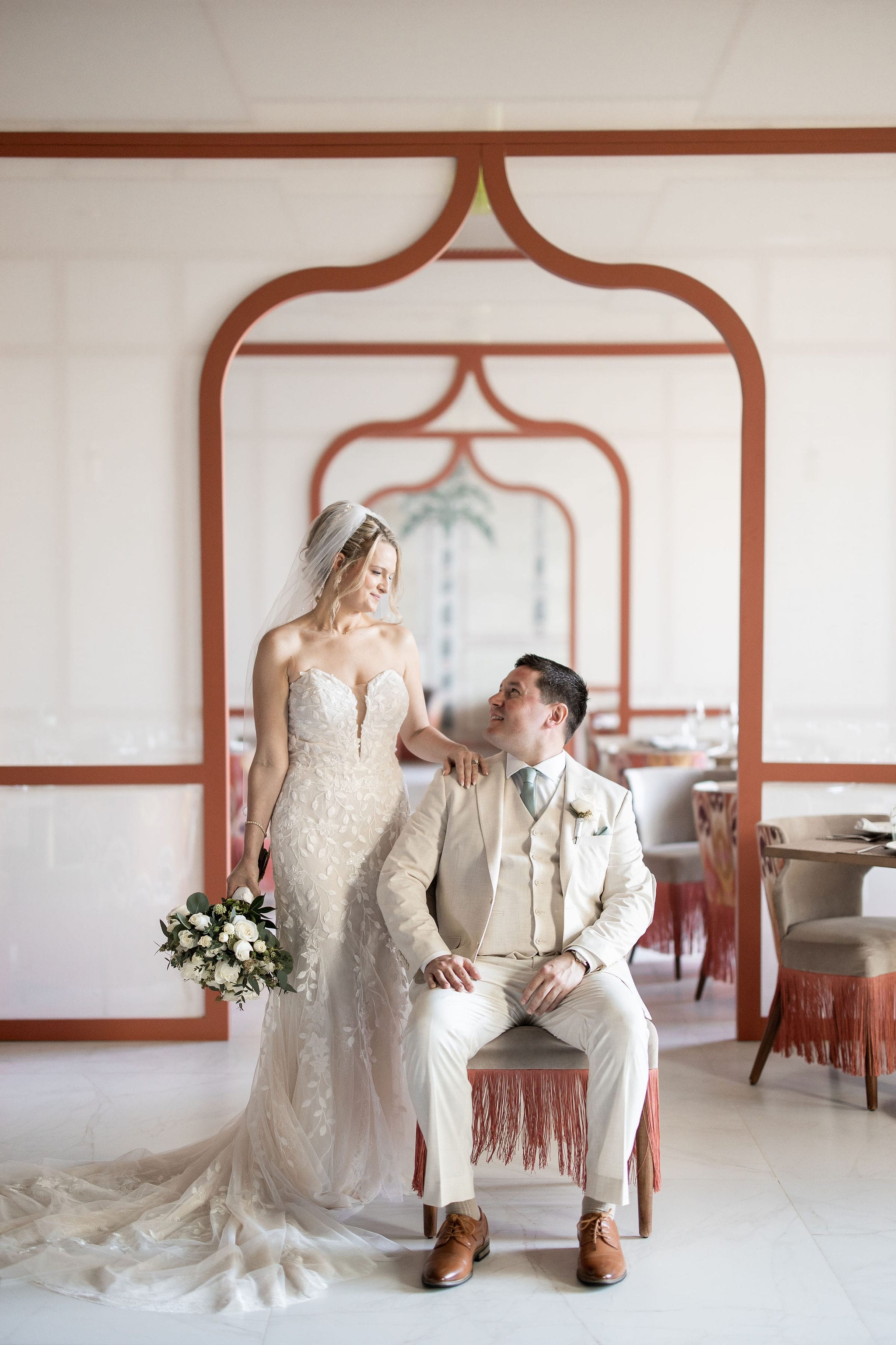Lauren and Ben at their wedding; she stands with bouquet beside groom seated indoors in formal attire.