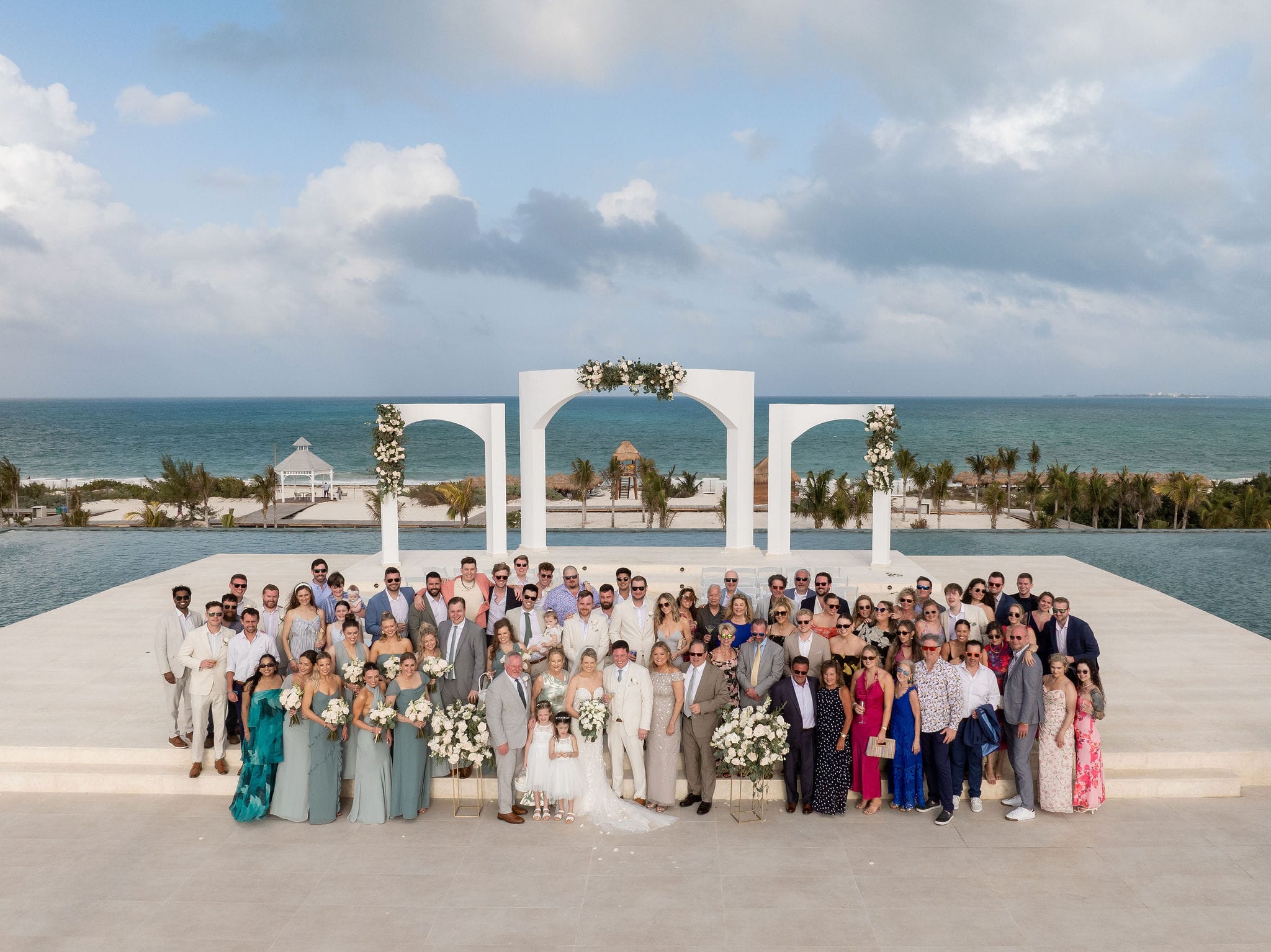 Lauren and Ben&rsquo;s wedding party poses outdoors by the ocean beneath a floral white arch at a destination wedding.