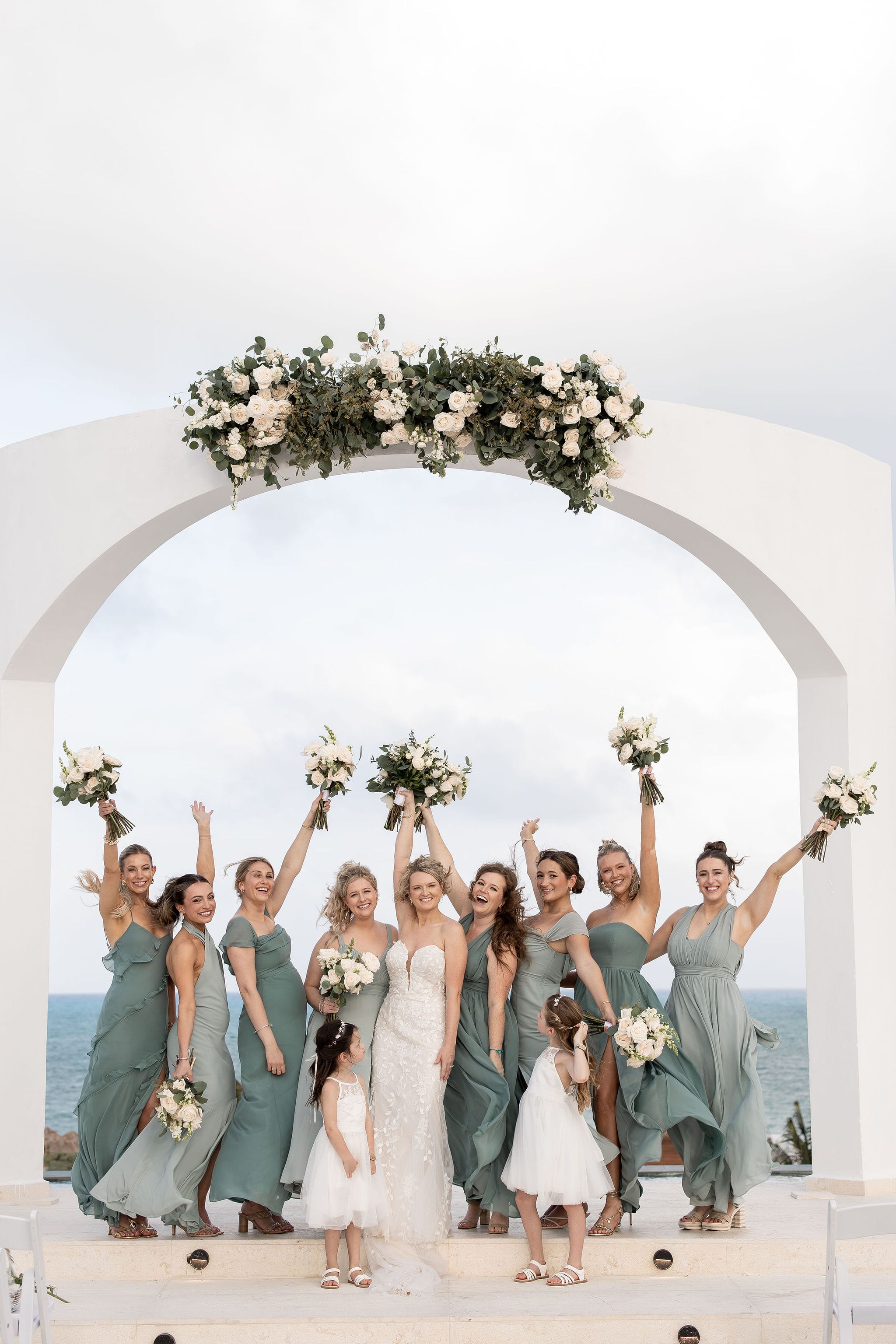 Lauren and Ben&rsquo;s destination wedding: bride, bridesmaids, and flower girls beneath a seaside floral arch.