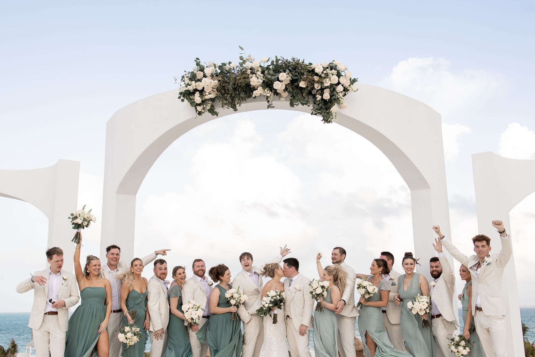 Lauren and Ben pose beneath a white floral arch by the ocean at their destination wedding.