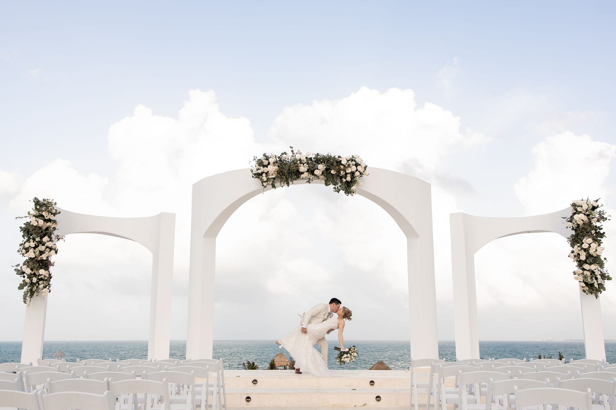 Lauren and Ben share a kiss beneath floral white arches by the ocean at their destination wedding.