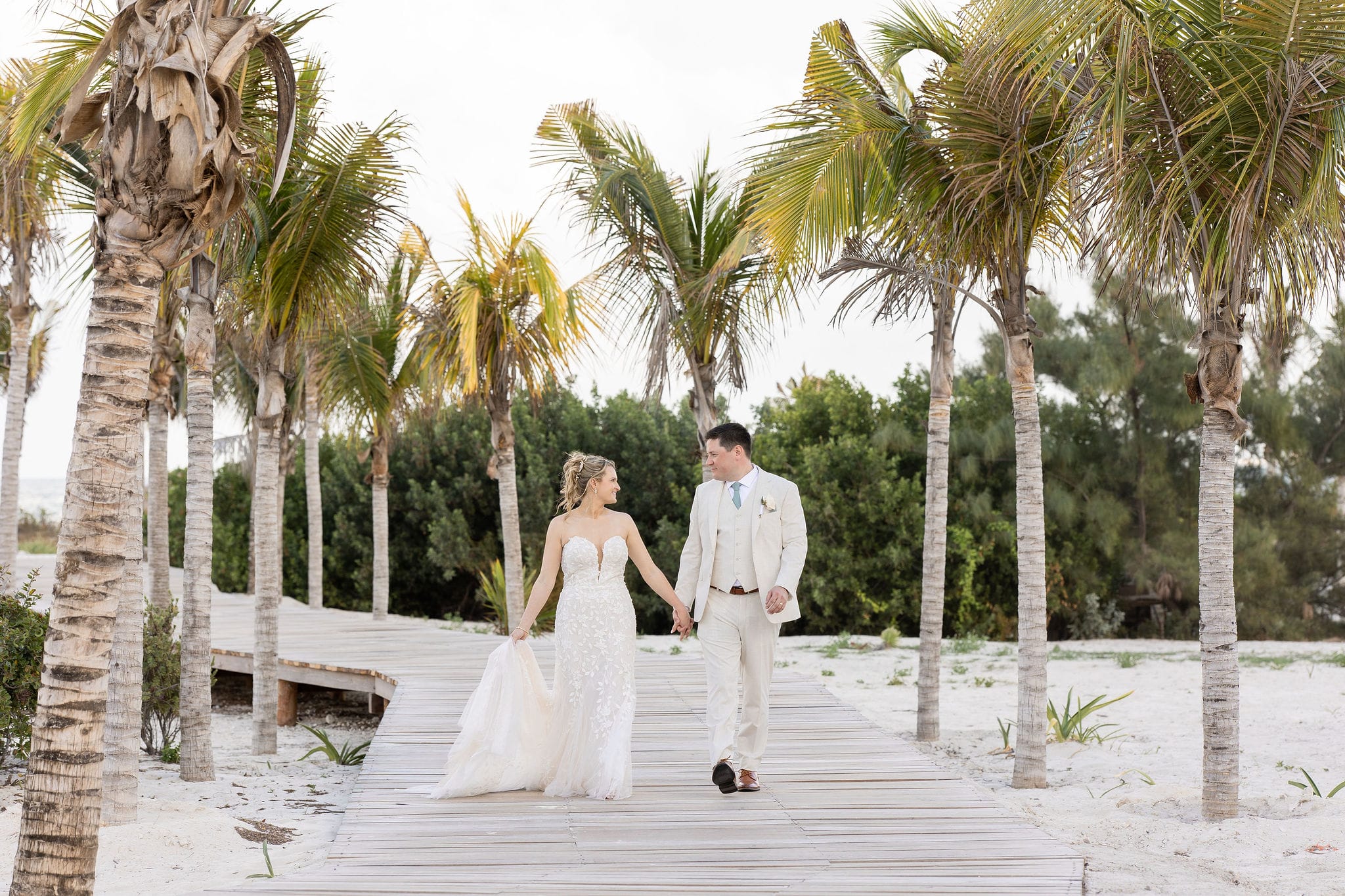 Lauren and Ben walk hand in hand on a wooden path lined with palm trees at their destination wedding.