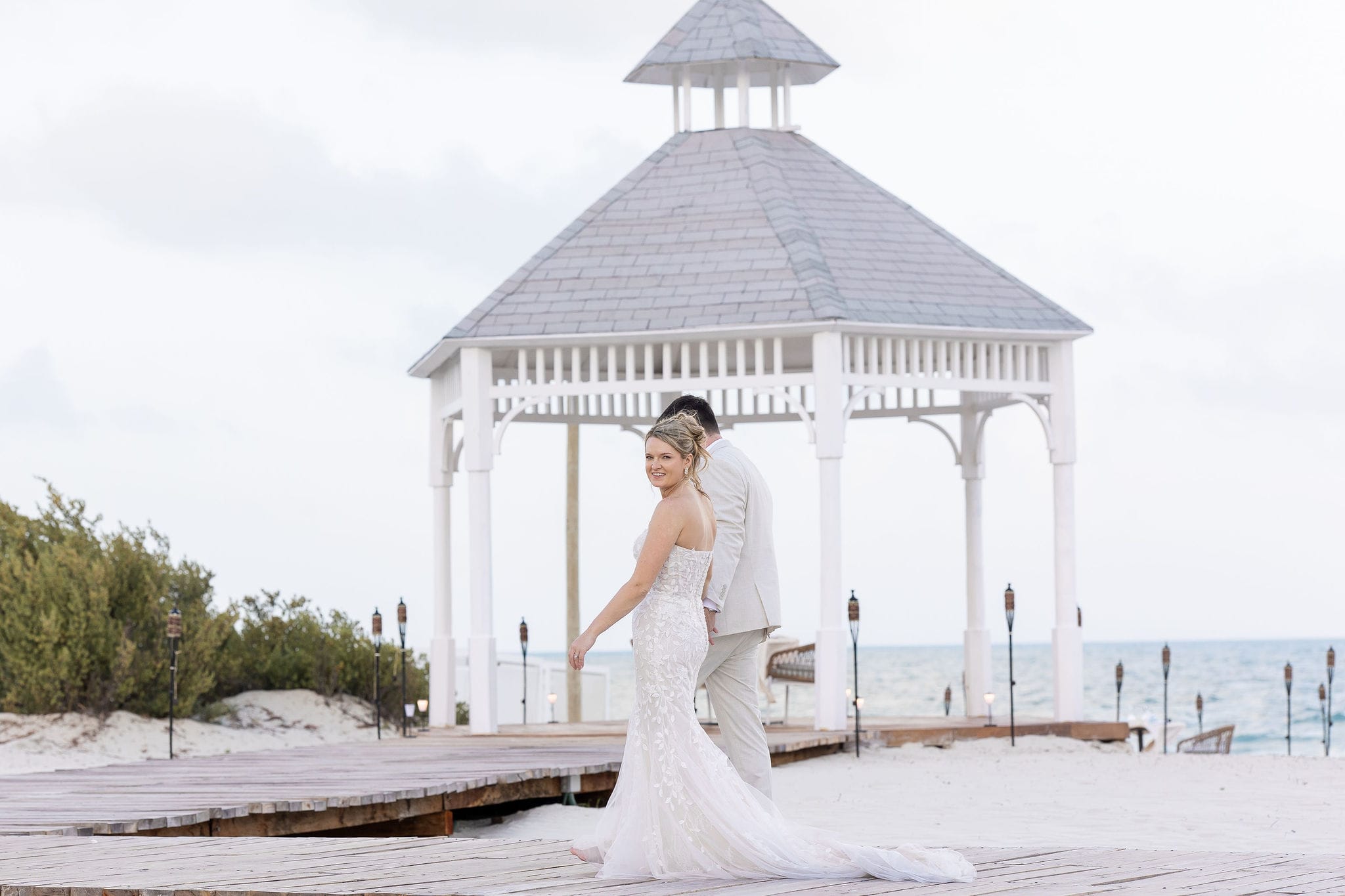 Lauren and Ben stand on a boardwalk by a white gazebo, celebrating their destination wedding at the beach.
