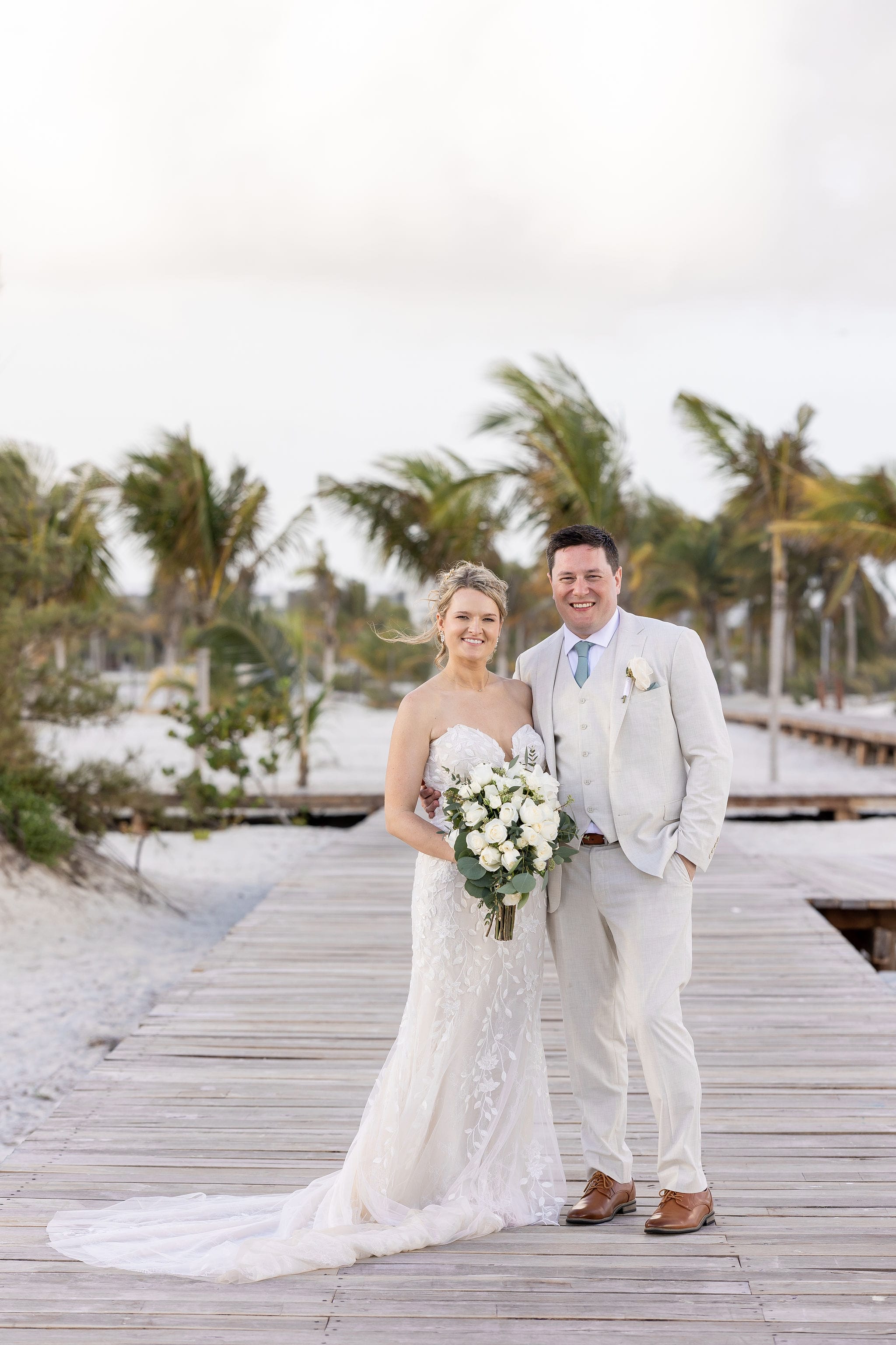 Lauren and Ben pose on a wooden boardwalk at a destination wedding beach with palm trees behind them.