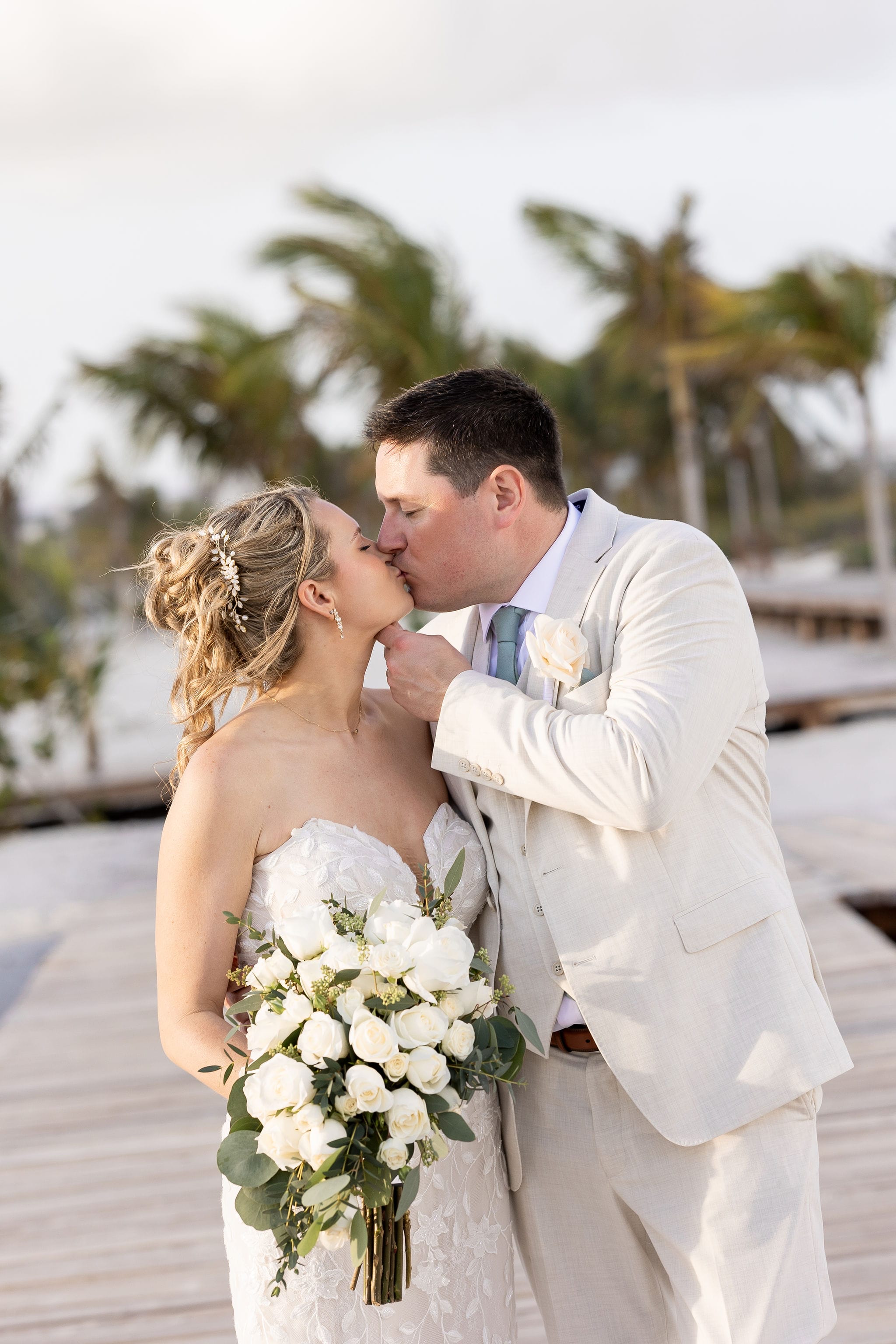 Lauren and Ben share a kiss on a boardwalk at their destination wedding, holding white roses, palm trees behind.