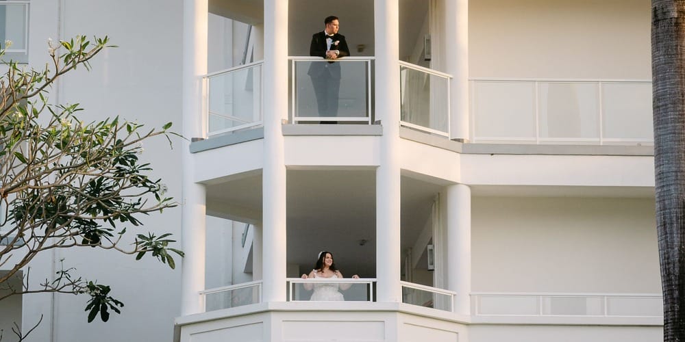 Andrea stands on an upper balcony and Kevin on a lower one at a white building for their destination wedding.