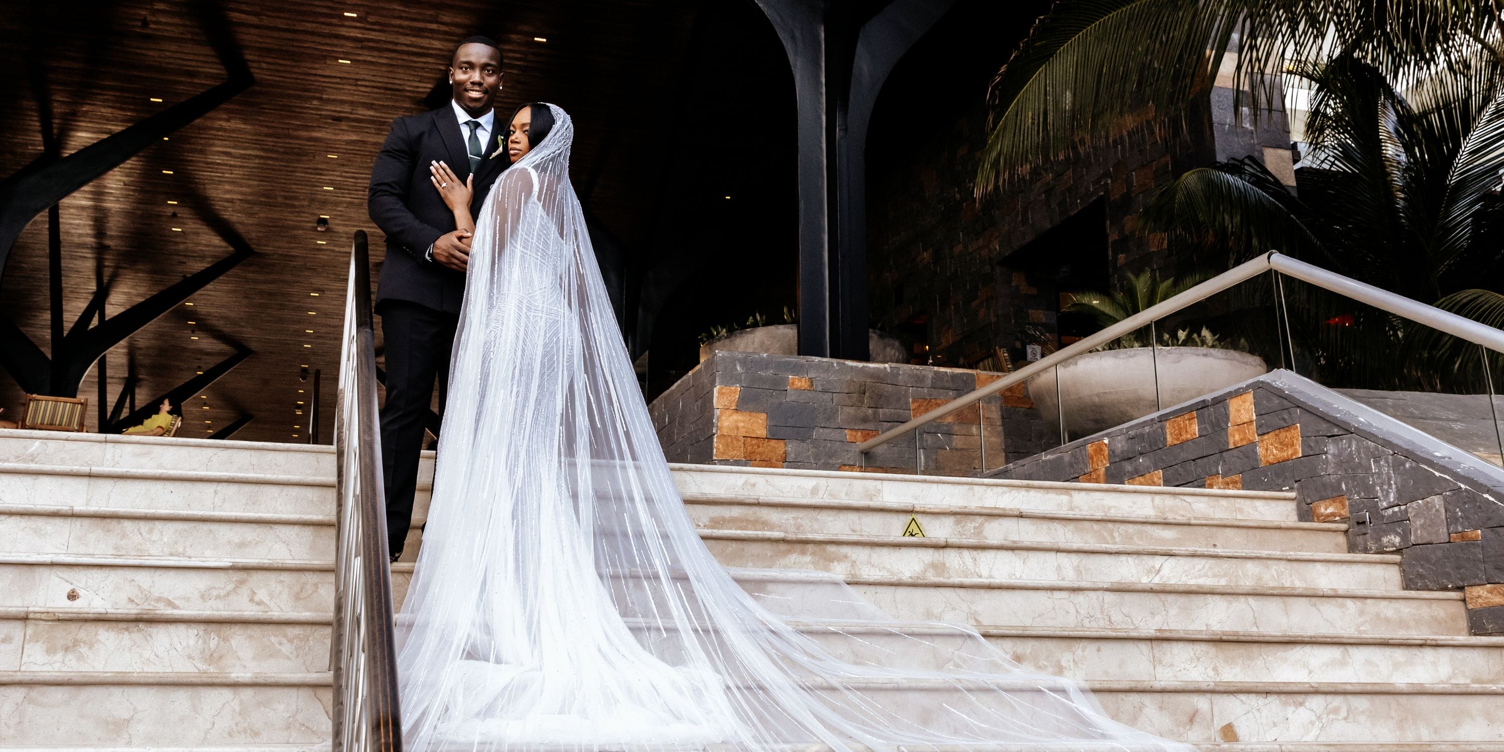 Quishona and Delon pose on stairs at their destination wedding, bride in veil, modern building behind.