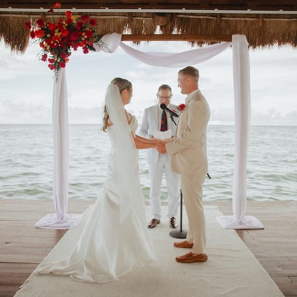 Couple exchange vows at their destination wedding under a floral arch by the water, officiant present.