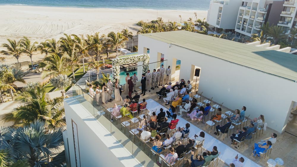 A destination wedding ceremony is held on a rooftop above the beach, with guests seated outdoors.