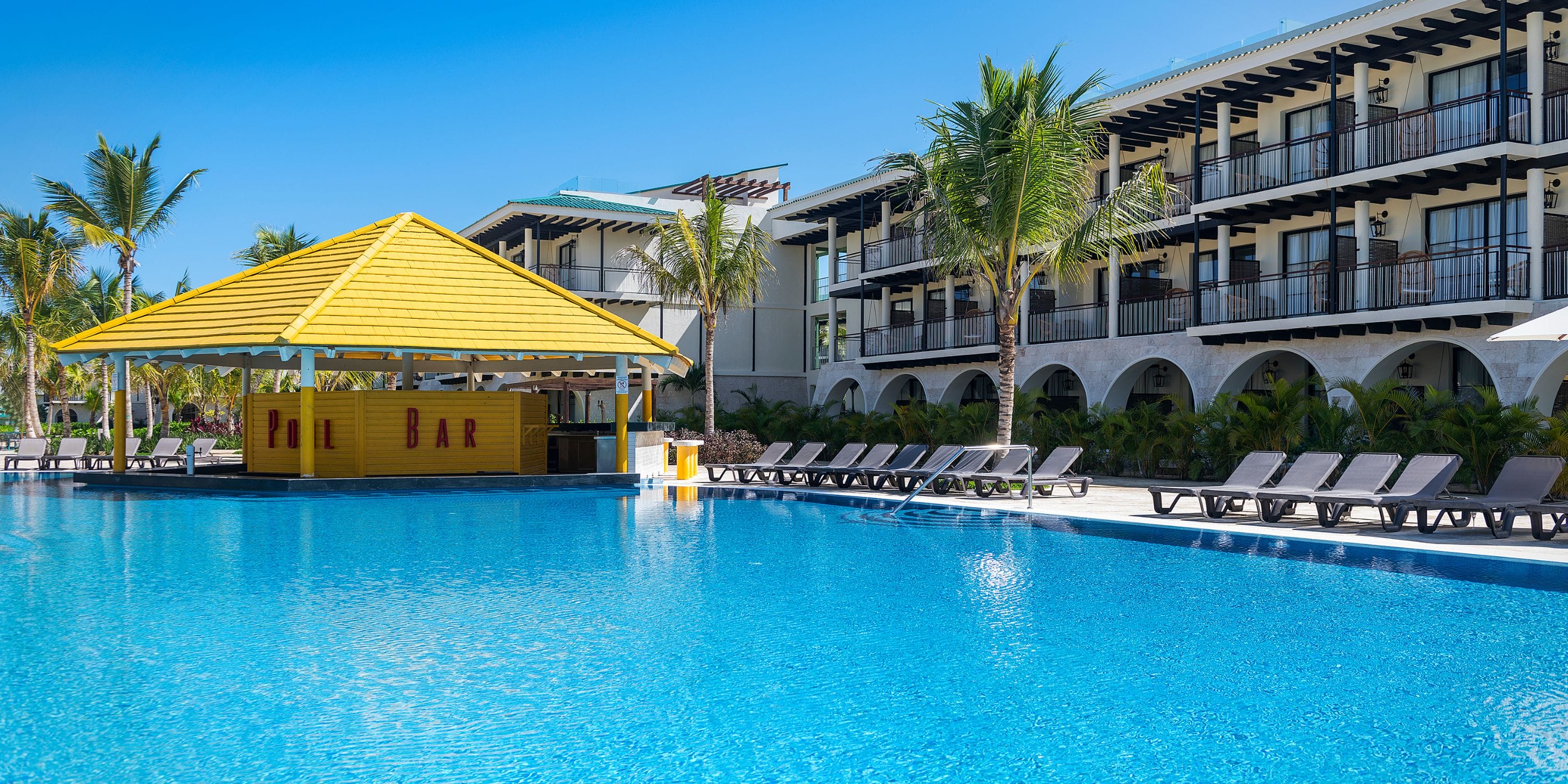 Spacious pool with lounge chairs, yellow bar, and palm trees&mdash;ideal for a destination wedding at El Beso.