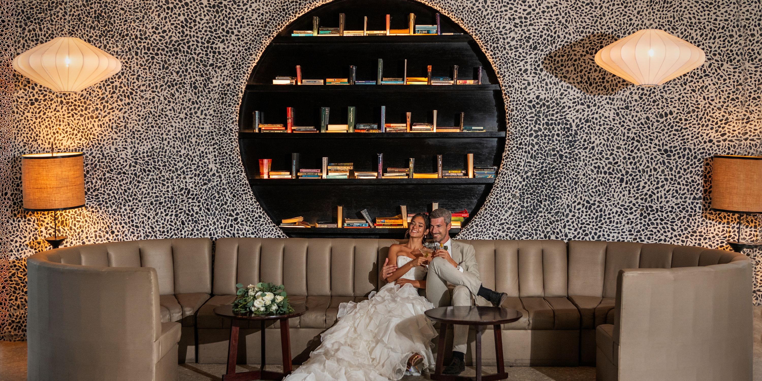 At their destination wedding, bride and groom sit on a curved sofa before a circular bookshelf.