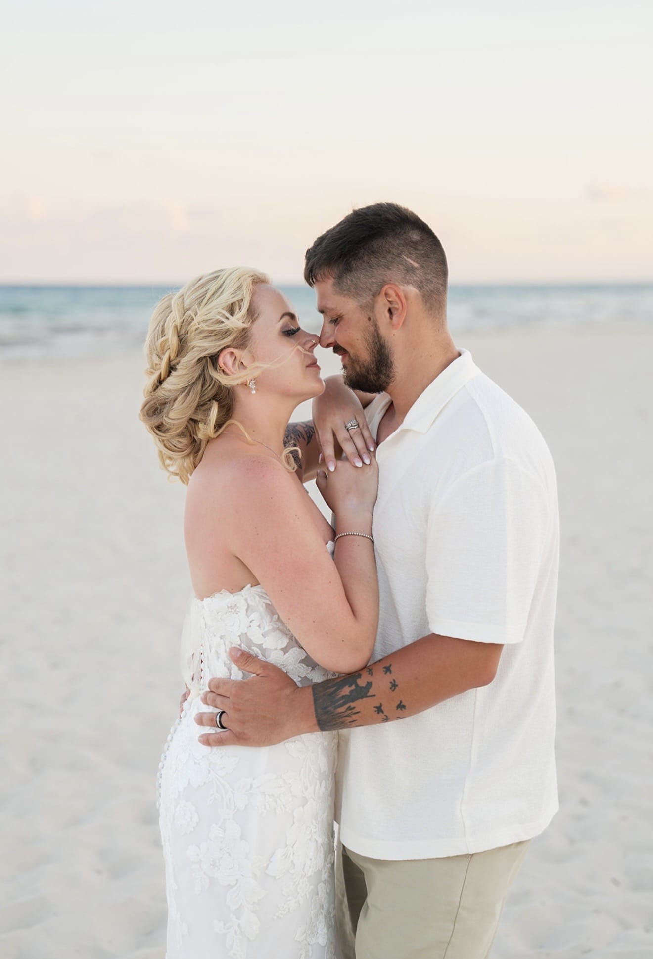 Hailey and Spencer embrace on a sandy beach at sunset during their destination wedding, facing closely.
