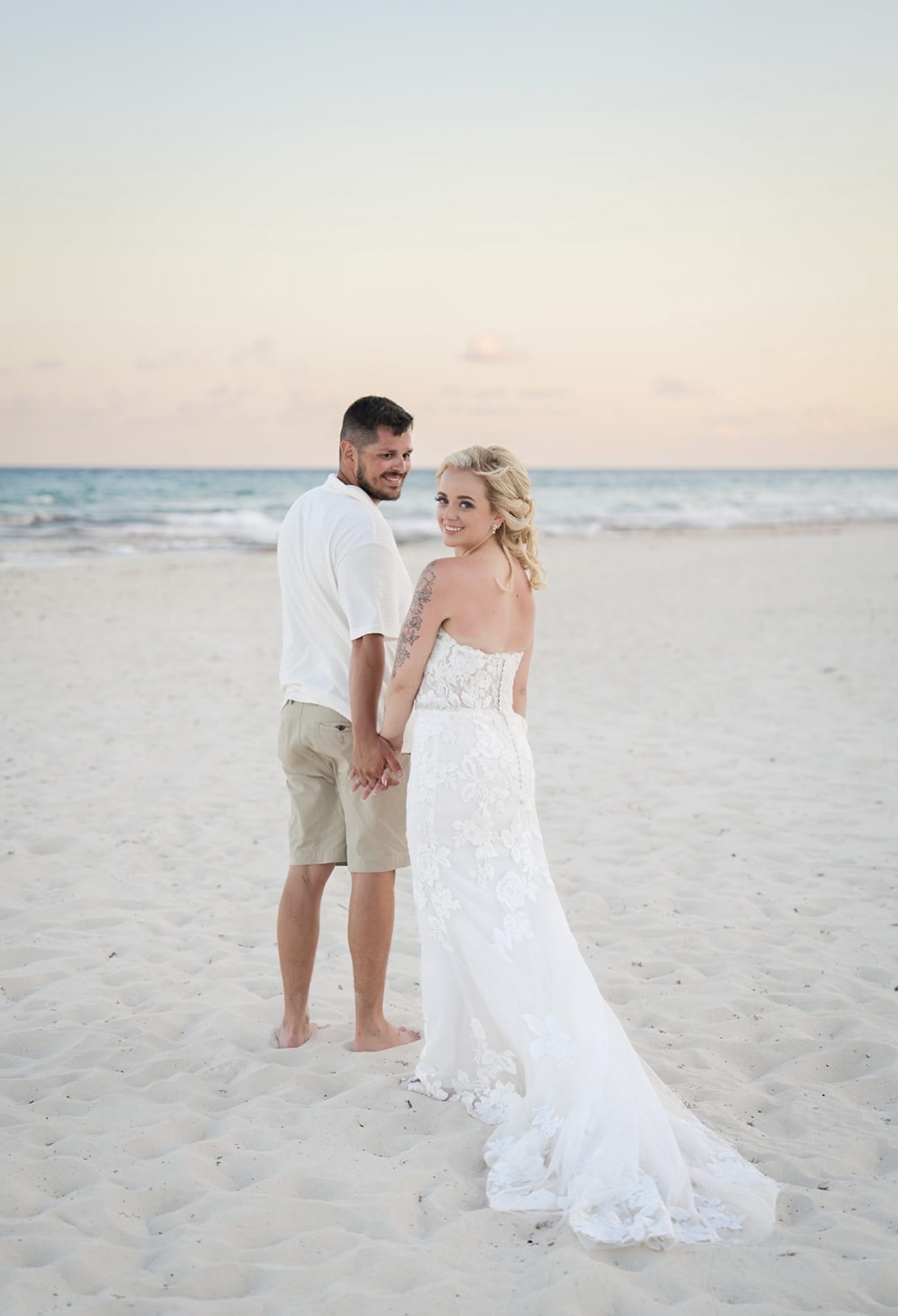 Hailey and Spencer hold hands on a sandy beach during their destination wedding, she in a white dress.
