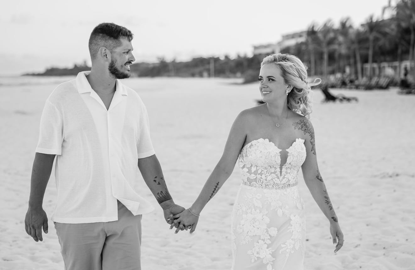 Hailey and Spencer stroll hand-in-hand on a sandy beach, sharing a loving look at their destination wedding.