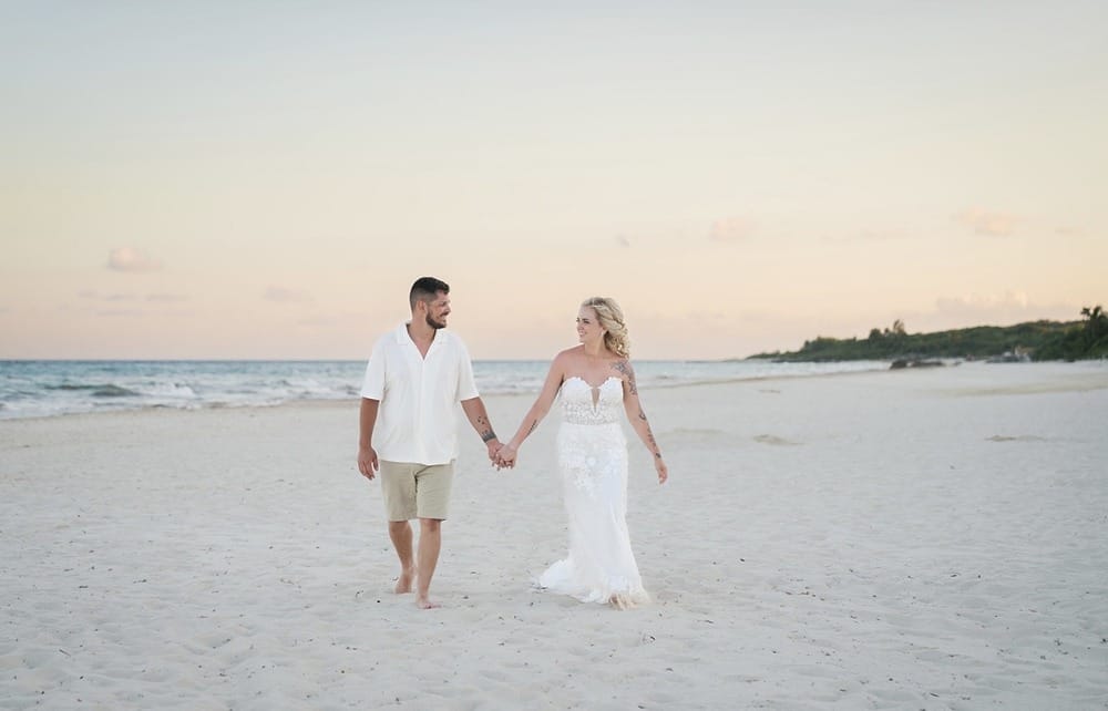 Hailey and Spencer stroll hand in hand on a sandy beach at sunset, dressed for their destination wedding.