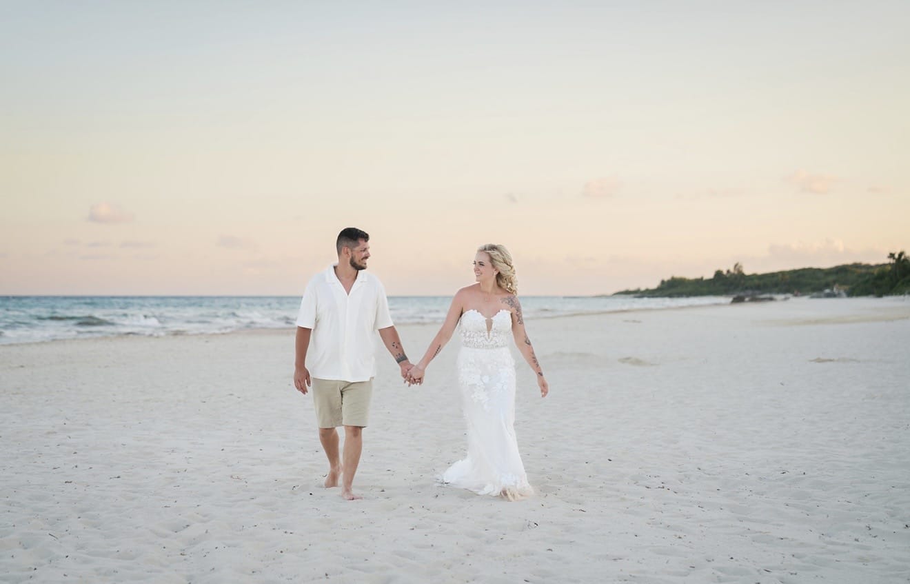 Hailey and Spencer stroll hand in hand on a sandy beach at sunset, dressed for their destination wedding.